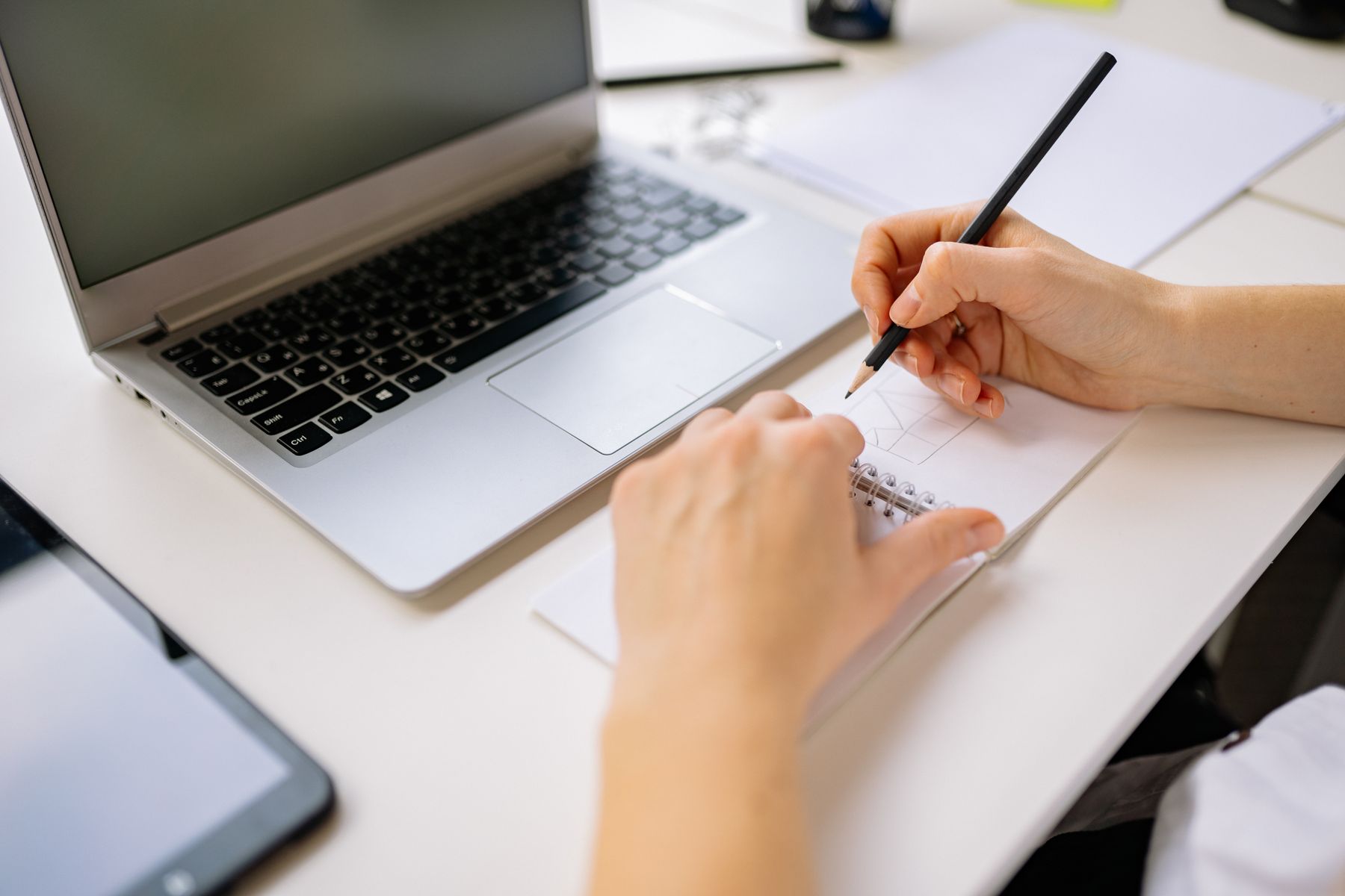 Hands sketching ideas in a notebook beside a laptop